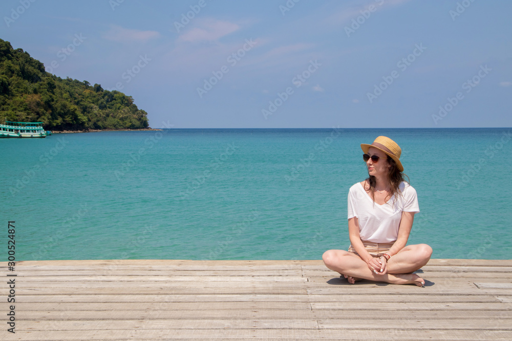 Young woman in a hat sits on a pier near the clear turquoise sea