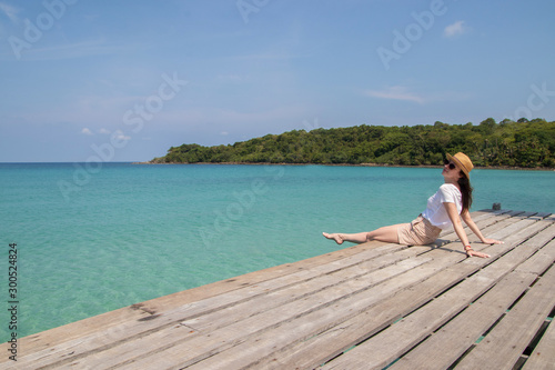 Young woman in a hat sits on a pier near the clear turquoise sea