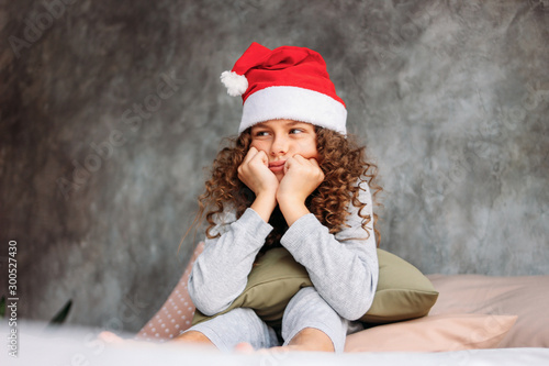 Curly haired beautiful uhappy tween girl in Santa hat and pajamas sitting on bed with pillow, christmas morning time photo