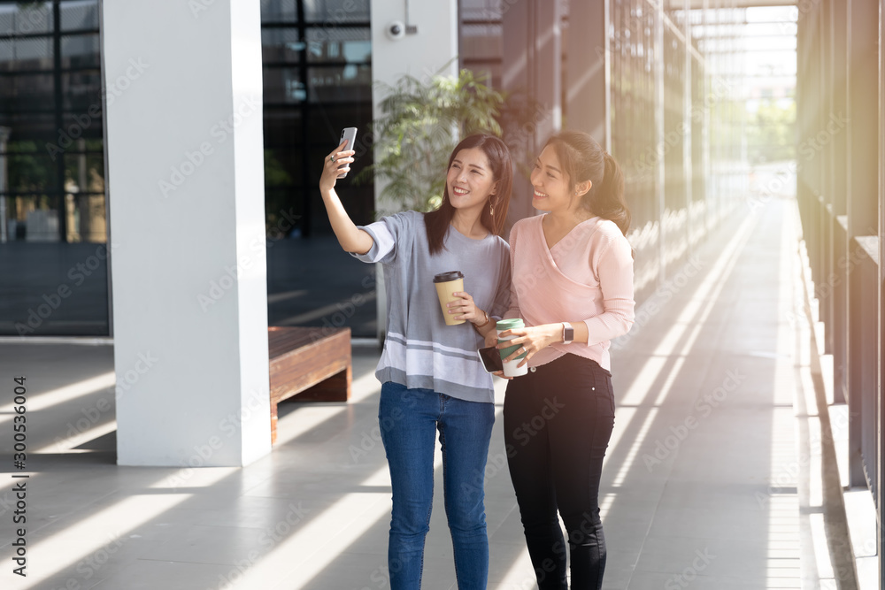 Cheerful young Asian woman in casual clothes two people selfie with smartphone and drinking take away coffee in paper cup in modern office. Technology and lifestyle concept. Cheerful young Asian woman in casual clothes two people selfie with smartphone and drinking take away coffee in paper cup in modern office. Technology and lifestyle concept.