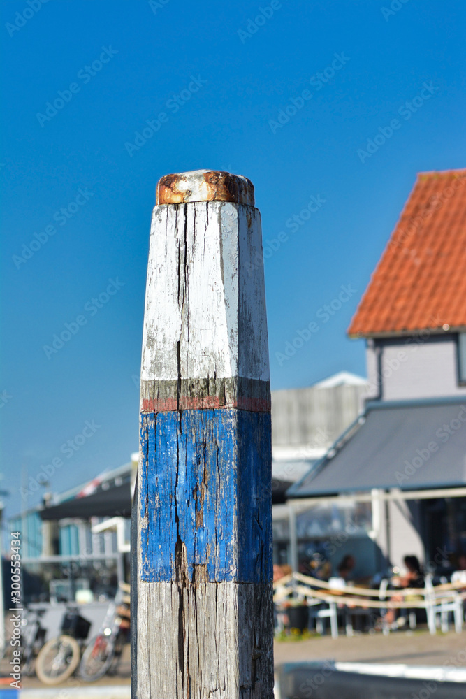 Wooden pole with old chipped blue and white paint for fastening boats at Oudeschild harbor in front of blue background