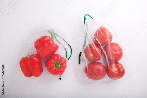 Bell peppers and tomatoes in textile grocery mesh bags. Red fruits and vegetables in reusable eco friendly packaging on white background.