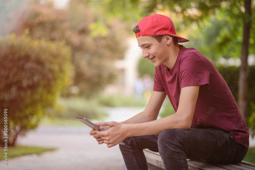 Teenager spends time in social networks. Smiling teenage boy with a tablet in the park. Handsome young man works on a tablet outdoors. Cheerful guy holds a tablet on the knees and looking to screen. Teenager spends time in social networks. Smiling teenage boy with a tablet in the park. Handsome young man works on a tablet outdoors. Cheerful guy holds a tablet on the knees and looking to screen.