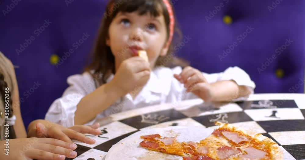 Children eat pizza. Two cute girls of four and seven years old are sitting at a table in a pizzeria holding slices of pizza and eating. Slow motion 50 FPS 4k. Close up portrait Children eat pizza. Two cute girls of four and seven years old are sitting at a table in a pizzeria holding slices of pizza and eating. Slow motion 50 FPS 4k. Close up portrait