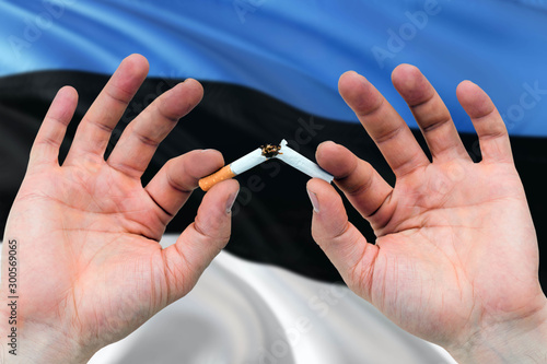 Estonia quit smoking cigarettes concept. Adult man hands breaking cigarette. National health theme and country flag background.