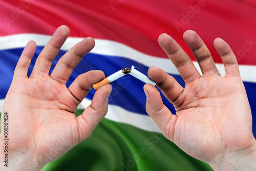 Gambia quit smoking cigarettes concept. Adult man hands breaking cigarette. National health theme and country flag background.