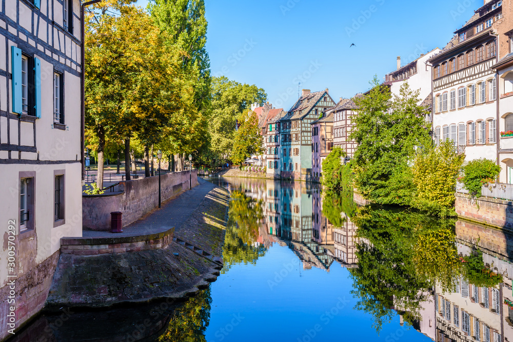 The river Ill canal bathes the Petite France quarter in Strasbourg, France, lined with half-timbered houses reflecting in the still waters under a bright sunshine.