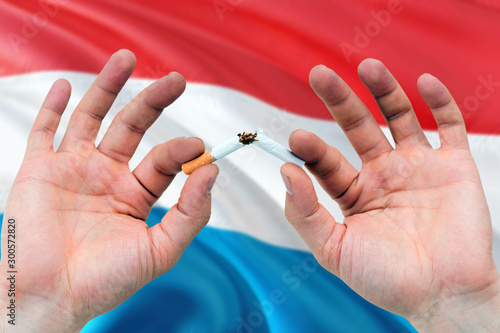 Luxembourg quit smoking cigarettes concept. Adult man hands breaking cigarette. National health theme and country flag background.