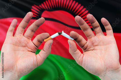 Malawi quit smoking cigarettes concept. Adult man hands breaking cigarette. National health theme and country flag background.