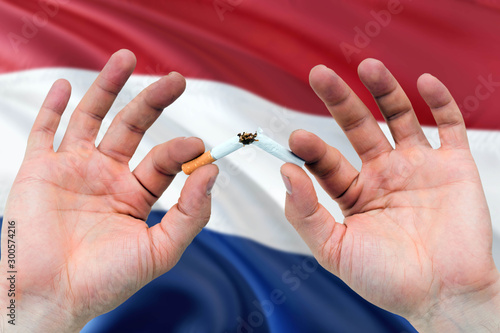 Netherlands quit smoking cigarettes concept. Adult man hands breaking cigarette. National health theme and country flag background.