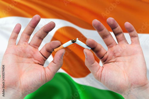 Niger quit smoking cigarettes concept. Adult man hands breaking cigarette. National health theme and country flag background.