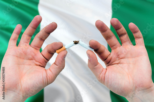 Nigeria quit smoking cigarettes concept. Adult man hands breaking cigarette. National health theme and country flag background.
