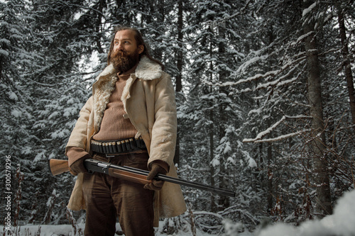 Portrait of brutal hunter man in winter vintage clothes with beard in forest with gun