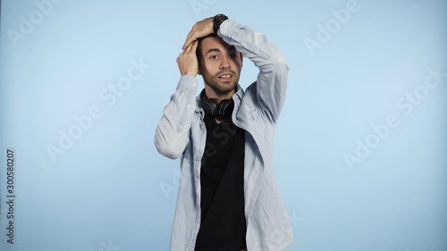 Footage over blue background of a caucasian young man that keep combing his dark short hair and stares at the camera. Feel unconfident and shy. Guy has black headphones on his neck photo