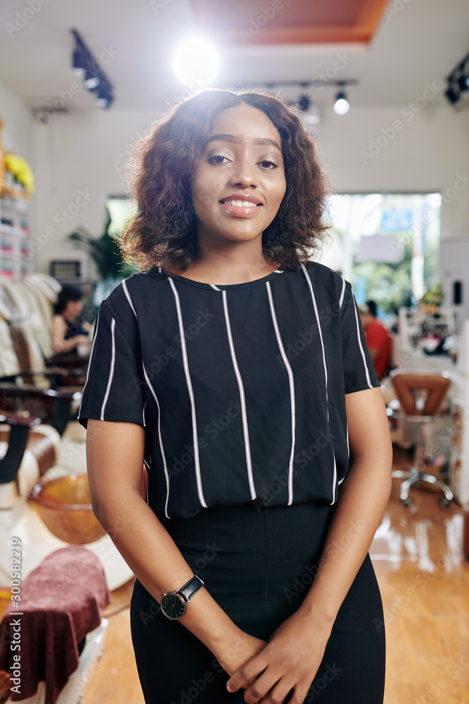 Portrait of young smiling Black woman standing in beauty salon where she works as a hairdresser Portrait of young smiling Black woman standing in beauty salon where she works as a hairdresser