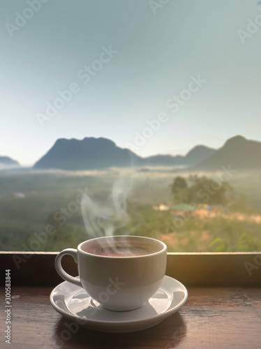 White ceramic coffee cup on wooden table in morning with sunlight over blurred mountains landscape 