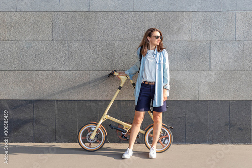 Fotografia One beautiful woman stands near a stone wall and holds a folding bike, looks at the sun