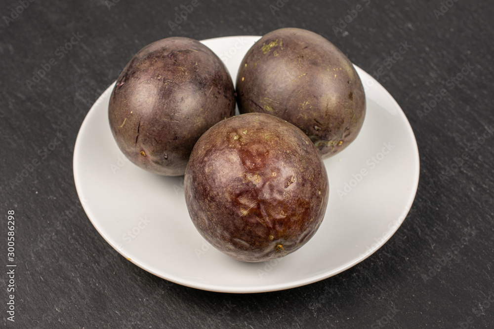 Group of three whole sweet dark purple passion fruit on white ceramic plate on grey stone Group of three whole sweet dark purple passion fruit on white ceramic plate on grey stone
