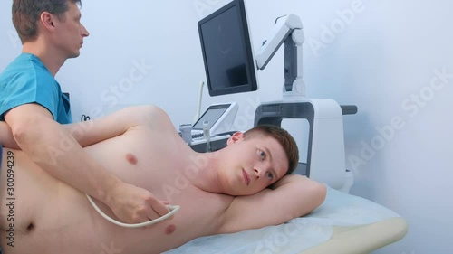 Echocardiography. Man doctor examining guy patient's heart by using ultrasound equipment. He runs ultrasound sensor over man's chest on heart area, working on scanner panel, looking at screen. photo