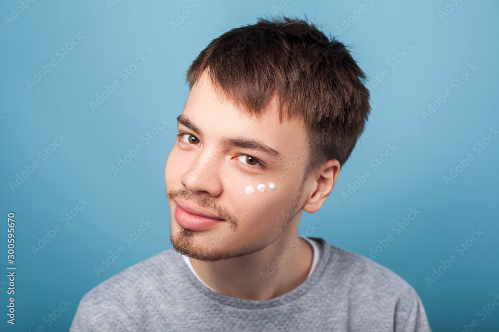 Fototapeta premium Skincare routine. Portrait of cheerful brunette man with small beard smiling at camera, having spots of moisturizer cream under eyes, male facial cosmetics. studio shot isolated on blue background