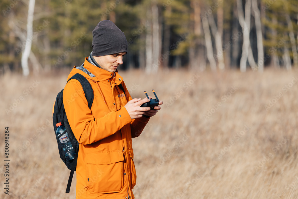 Fototapeta premium A man in an orange jacket, with a backpack and a hat, controls a quadrocopter drone outdoors in autumn.