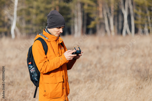 A man in an orange jacket, with a backpack and a hat, controls a quadrocopter drone outdoors in autumn.