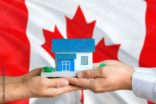 Canada real estate concept. Man and woman holding miniature house in hands. Citizenship theme and national flag on background.