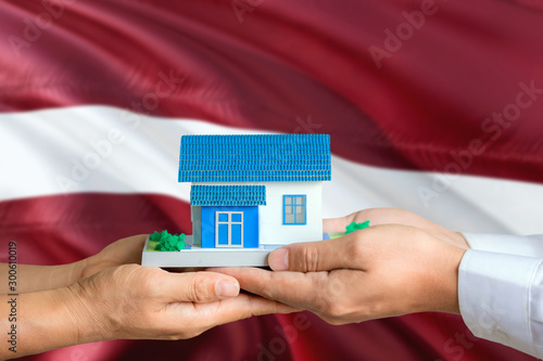 Latvia real estate concept. Man and woman holding miniature house in hands. Citizenship theme and national flag on background.