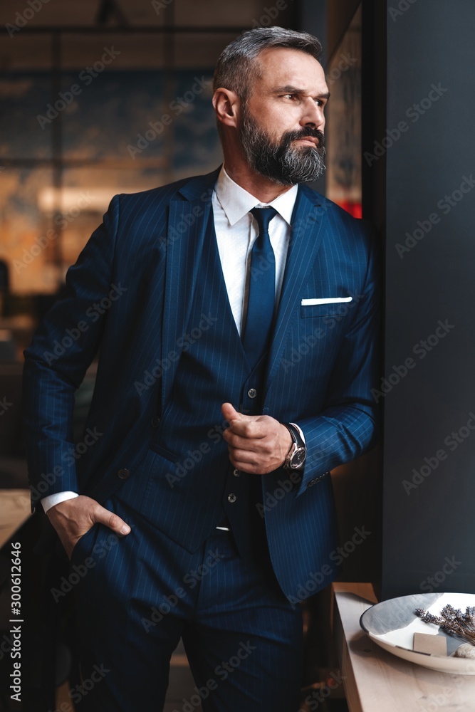 Stylish bearded man in a suit standing in modern office - obrazy, fototapety, plakaty 