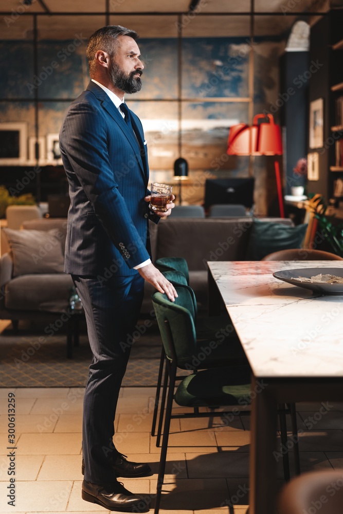 Mature handsome businessman stand and hold glass of whiskey in his own office. Mature handsome businessman stand and hold glass of whiskey in his own office.