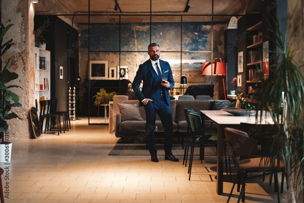 Mature handsome businessman stand and hold glass of whiskey in his own office. Mature handsome businessman stand and hold glass of whiskey in his own office.