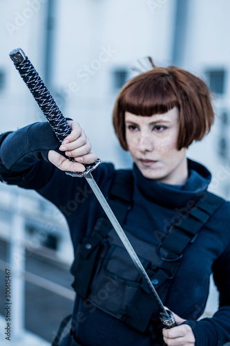 portrait of young redhead woman with japanese sword dressed in futuristic cyberpunk costume standing on the roof. 