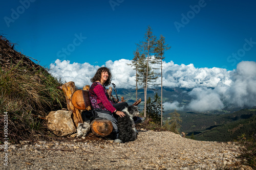 Woman and dog sitting on a bench resting while hiking to the Tschaffonhuette on a high mountain with white clouds above a valley in the background photo