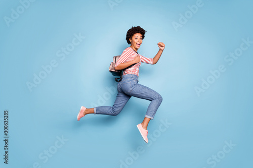Full length photo of cheerful funny teenage afro american girl jump run college hold backpack wear striped shirt denim jeans pink sneakers isolated over blue color background photo