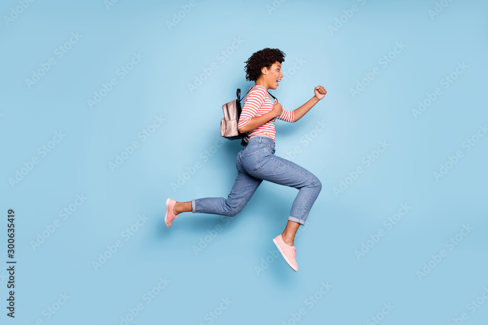 Full body profile side photo of positive cheerful afro american girl jump run fast summer school sales hold bag wear striped shirt denim jeans isolated over blue color background