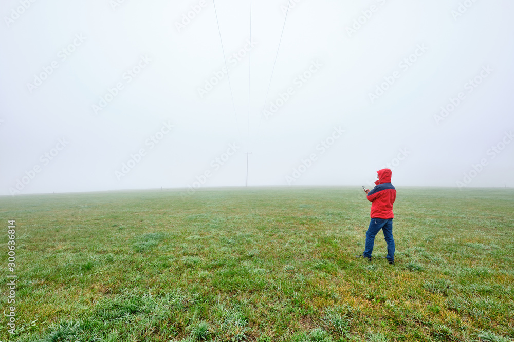 Rear view of man in red jacket standing on a green meadow and looking on his smartphone in front of a foggy nowhere landscape. Seen in October in Germany, Bavaria near Oedenberg. Rear view of man in red jacket standing on a green meadow and looking on his smartphone in front of a foggy nowhere landscape. Seen in October in Germany, Bavaria near Oedenberg.