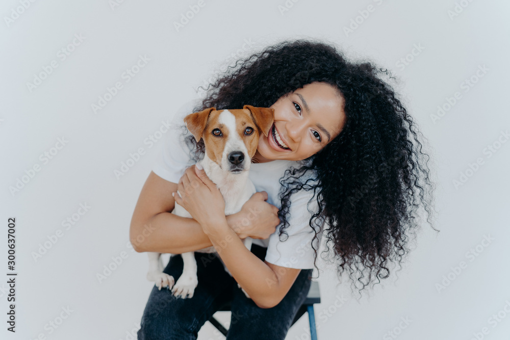 Happy curly woman tilts head, holds pedigree dog, has cheerful expression, smiles pleasantly, has curly hairstyle, wears white t shirt, isolated. Afro lady petting favourite domestic animal. Happy curly woman tilts head, holds pedigree dog, has cheerful expression, smiles pleasantly, has curly hairstyle, wears white t shirt, isolated. Afro lady petting favourite domestic animal.