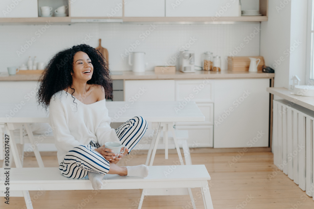 Horizontal shot of overjoyed dark skinned woman laughs pleasantly, drinks coffee, looks out of window in kitchen, dressed in fashionable clothes. Smiling lady with hot tasty beverage, relaxes at home Horizontal shot of overjoyed dark skinned woman laughs pleasantly, drinks coffee, looks out of window in kitchen, dressed in fashionable clothes. Smiling lady with hot tasty beverage, relaxes at home