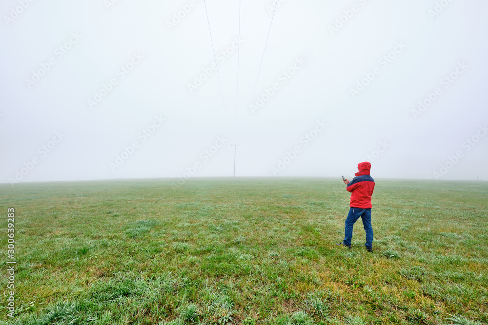Rear view of man in red jacket standing on a green meadow and looking on his smartphone in front of a foggy nowhere landscape. Seen in October in Germany, Bavaria near Oedenberg.