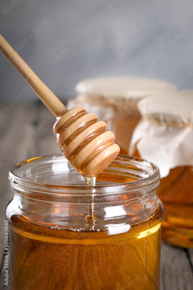 Pouring aromatic honey into jar, closeup. Honey in glass jars and honeycombs wax on wooden background. Wooden stick , instruments Pouring aromatic honey into jar, closeup. Honey in glass jars and honeycombs wax on wooden background. Wooden stick , instruments