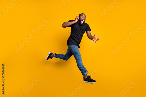 Wait i hurry. Full body profile side photo of crazy funny afro american guy jump speak cellphone run buy black friday discounts wear t-shirt denim jeans isolated yellow color background © deagreez