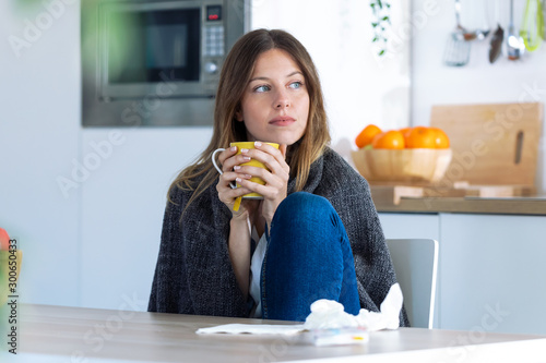 Sick and cold young woman drinking hot beverage while sitting in the kitchen at home.