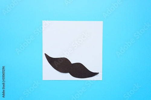 Black paper moustache and writing paper sticker on a blue background. The concept of awareness of men's health and prostate cancer. Space for text photo