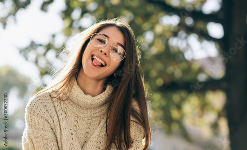 Close-up outdoor portrait of funny student woman showing tongue out, looking to the camera, wearing knitted sweater and transparent eyeglasses. Beautiful happy young female posing outside on sunlight