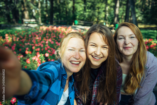 Happy young girls have fun laughing and taking selfies on a smartphone in the park on the background of green foliage and flowers. Blonde, brunette and girl with red hair, female friendship concept.