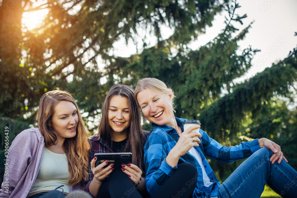 Happy young girls have fun laughing sitting on green grass in the park. Students in between lectures. Blonde, brunette and red-hair girl communicate, look at smartphone, concept of female friendship.