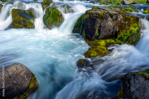 Waterfalls with clear blue water and green mossy boulders outdoors in the icelandic nature.