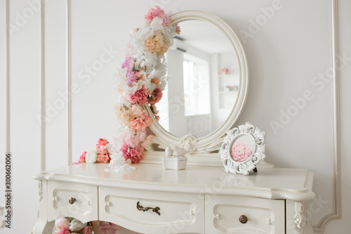 Vintage style boudoir table with round mirror and flowers. White bright room.