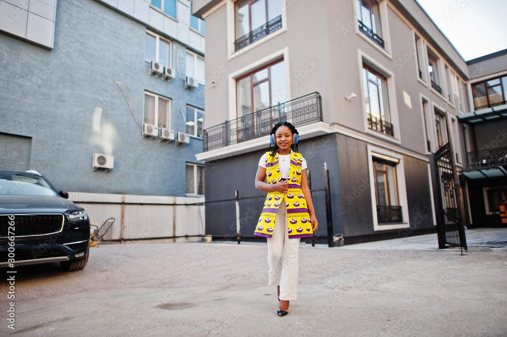 Stylish african american women in yellow jacket posed and earphones on street against modern building. Stylish african american women in yellow jacket posed and earphones on street against modern building.