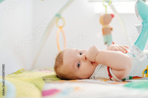 Fotografi Side view of beautiful six months old boy in colorful bodysuit looking at camera and sucking thumb while lying in bed surrounded by toys